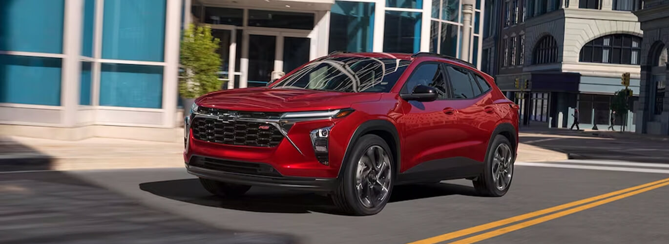 A red 2024 Chevrolet Trax RS driving through city streets. Experince the technology of the Trax today at York Chevy near, Bloomington, Indiana.